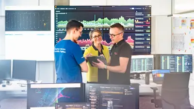 Three people discuss in front of large data screens in a modern office.