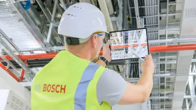 A Bosch technician wearing a hard hat and high-visibility vest uses a tablet for maintenance or inspection of complex wiring and installations in an industrial facility.